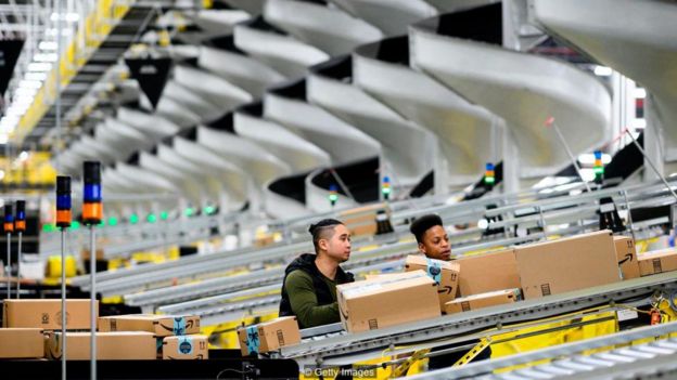 Dois homens trabalham em sistema operacional da Amazon, monitorando distribuição de caixas