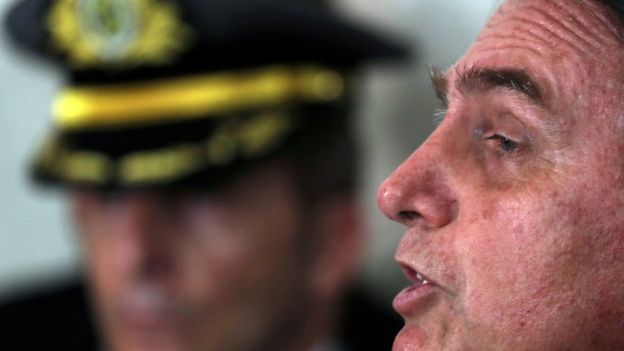 Bolsonaro aparece de perfil e falando, com homem fardado no plano de fundo