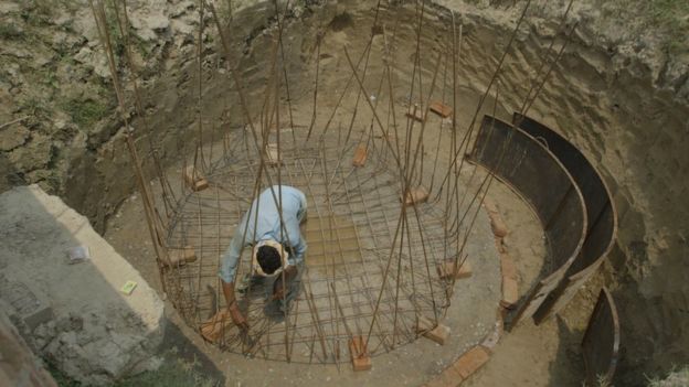 A man in a large hole constructing a biodigester with iron wires