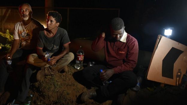 Three men sit by a road, holding water bottles