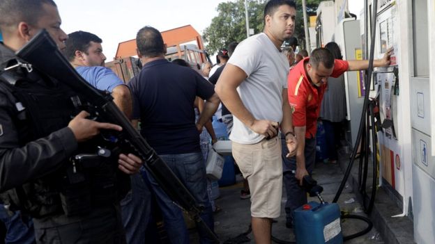 Policial armado patrulha posto de gasolina no Rio de Janeiro, enquanto pessoas enchem vasilhas com combustível