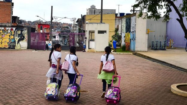 Três meninas com uniforme e mochilas caminham em praça pública