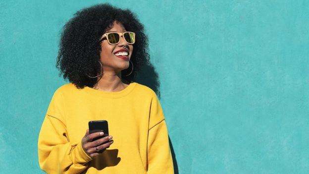 Jeune femme avec un pull jaune vif tenant un smartphone, posant devant un mur turquoise.