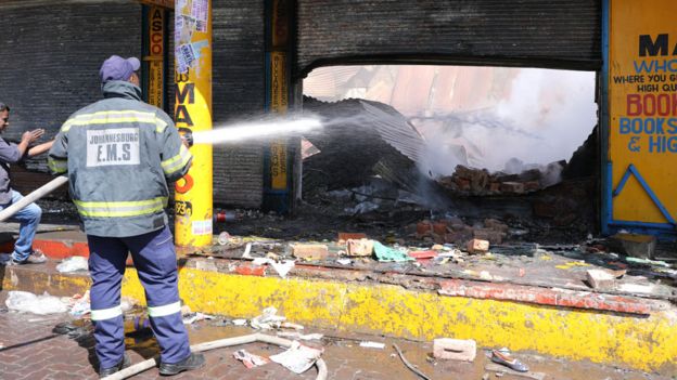 A shop keeper watches as a fireman douses down a burned and damaged property after overnight unrest and looting in Alexandra township, Johannesburg