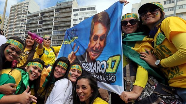 Mulheres com camisas verde amarelo seguram bandeira com rosto de Bolsonaro
