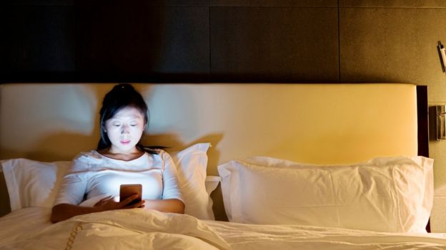 Mulher na cama, olhando para o smartphone em vez de dormir.