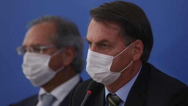 Sentados e de perfil, Paulo Guedes e Jair Bolsonaro de máscara