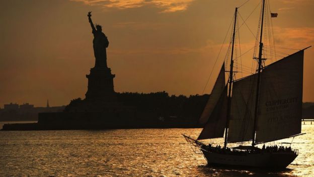 Vista de la estatua de la libertad al atardecer, con un barco antiguo de vela en la bahÃ­a
