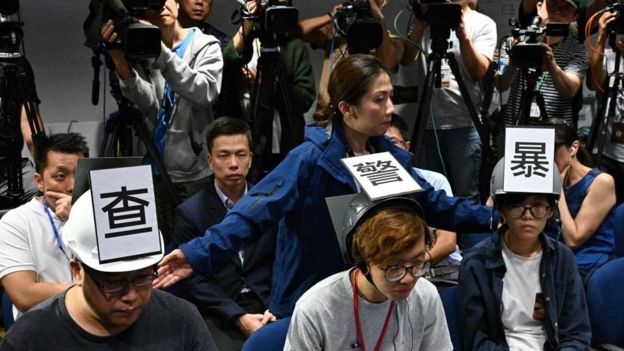 A police media relation officer (behind, C) stands behind six journalists (three not pictured), including Ronson Chan Ron-sing (L), a member of the executive committee of local media company Stand News, wearing protective press helmets with Chinese characters that collectively read Investigate Police Brutality Stop Police Lies in a press conference room at the police headquarters in Hong Kong on November 4, 2019,