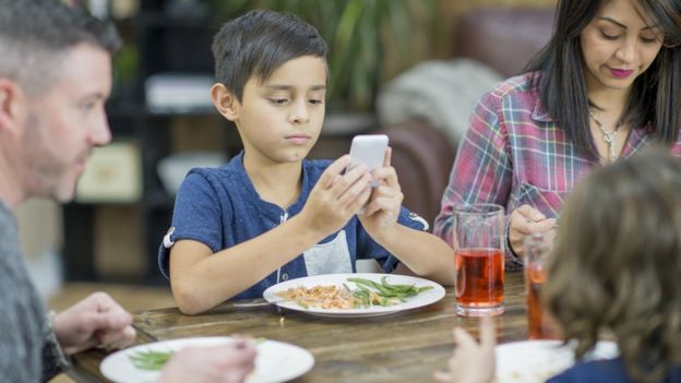 Um garoto segura seu celular em uma mesa durante a refeição