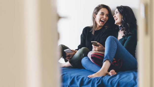 Duas adolescentes sentadas em uma cama, sorrindo com os celulares na mão