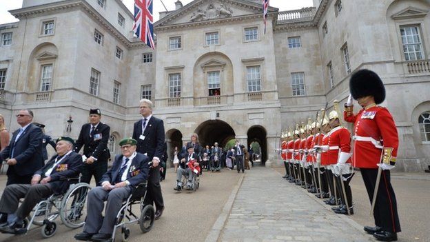 A guard of honour saluting WW2 veterans in wheelchairs as they being pushed by relatives and friends through Horse Guards Parade