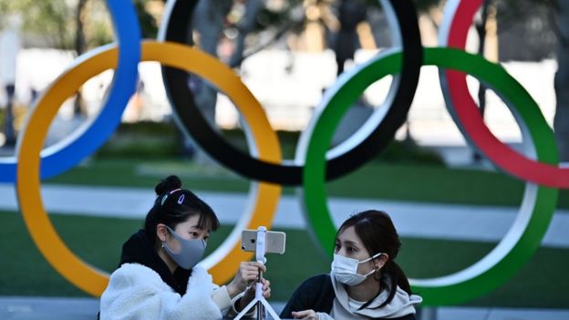 Mulheres japonesas com máscaras em frente a símbolo dos Jogos Olímpicos