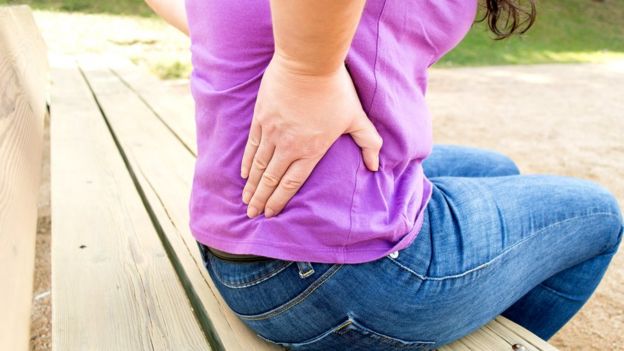 Mujer sentada en un banco con mano en la espalda en señal de dolor.