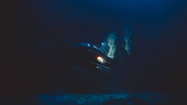 Submarino investiga as profundezas do Buraco Azul de Belize - imagem é toda escura, só é possível ver a luz do submarino iluminando pedaços de estalactites e refletindo de volta no equipamento