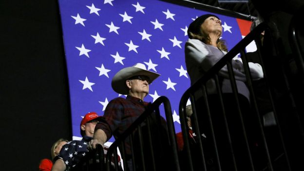 Apoiadores de Trump em fila durante evento, com bandeira dos EUA no plano de fundo