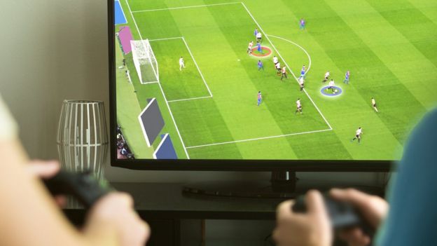 Una pantalla de televisión con un videojuego de fútbol y dos personas jugando.