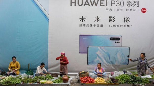 Women sell vegetables in front of a billboard advertising smartphones for China's Huawei Technologies Co., on June 1, 2019 in Mangshi, Yunnan Province, southwestern China