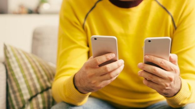 A man with a yellow jumper holding two identical smartphones