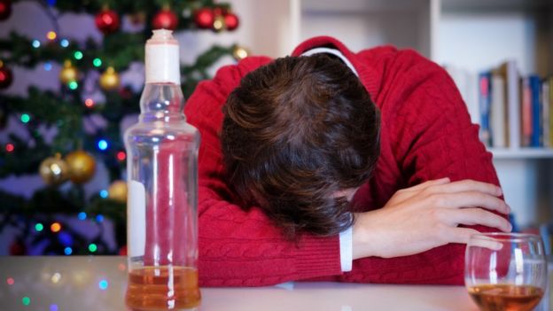Homem dormindo em cima da mesa com garrafa de bebida alcoolica na frente e árvore de natal atrás