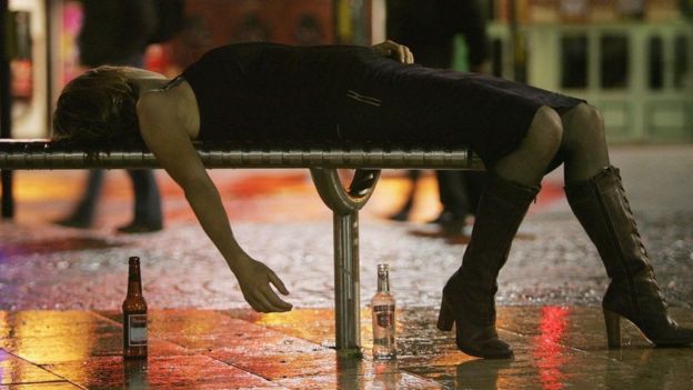 Una mujer recostada sobre un banco con botellas de cerveza en el suelo.
