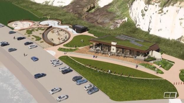 A digital impression of a restaurant and play park on a beach. To the left is a sandy play park, on the right is a brown restaurant with lots of glass panels and a grass roof. Th area sits at the base of white chalk cliffs in front of the sea.