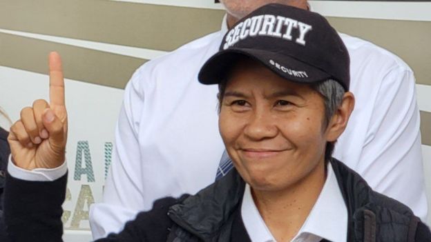Romana Didulo wearing black cap with word securityin white, holding up index finger.\