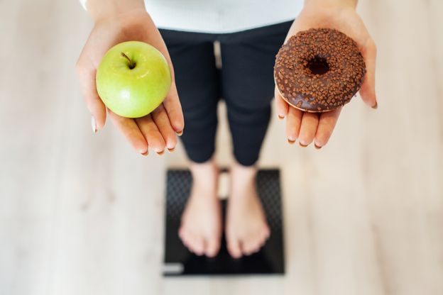 Imagem mostra mulher em pé sobre balança enquanto segura uma maçã e um donut.