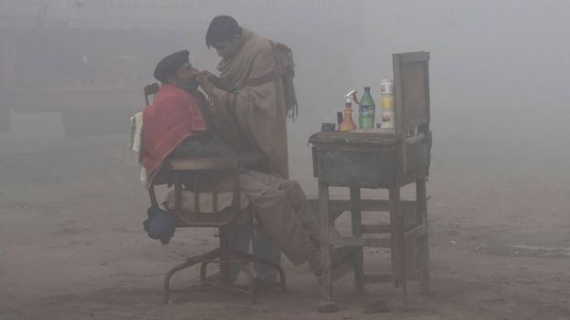 A Pakistani barber shaves a customer alongside a road amid heavy fog and smog conditions in Lahore on January 24, 2019.