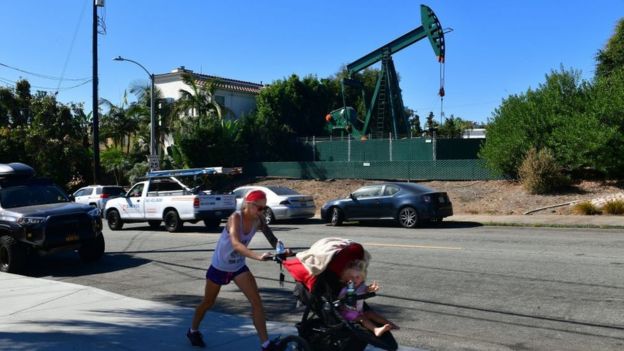 Una mujer camina con su bebé por una calle residencial de Los Ángeles y al fondo se ve un aparato extractor de petróleo.