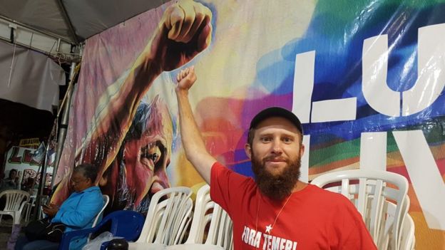 Inácio Sadovski sorri e levanta o braço em frente a cartaz com foto de Lula fazendo o mesmo gesto