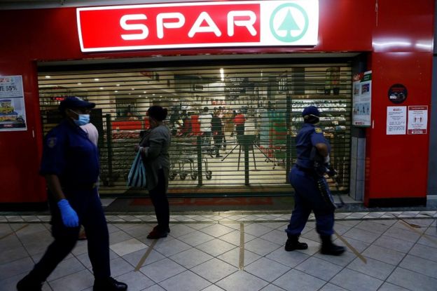 South African Police Service (SAPS) walk past shoppers queuing outside a food store during their operation in Sunnyside, Pretoria on April 7, 2020