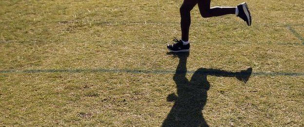 A runner trains on the grass and is shadowed by the sun
