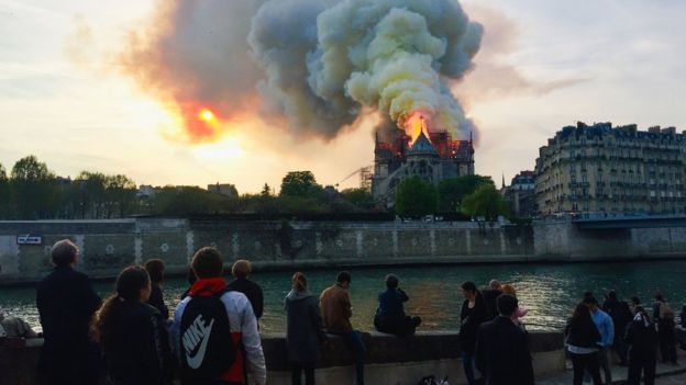 Nas margens do Rio Sena, pessoas observam incêndio da catedral do outro lado, durante entardecer