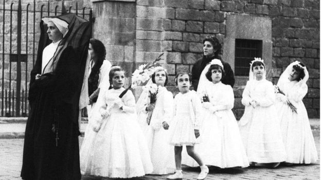 Una monja guía a un grupo de niñas vestidas de blanco para su primera comunión.