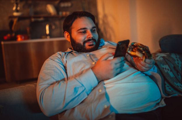 Un hombre obeso mirando su celular mientras come una hamburguesa