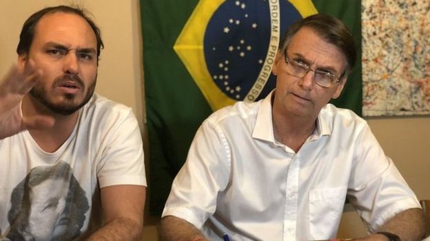Carlos Bolsonaro e Jair Bolsonaro sentados à mesa durante transmissão ao vivo de vídeo