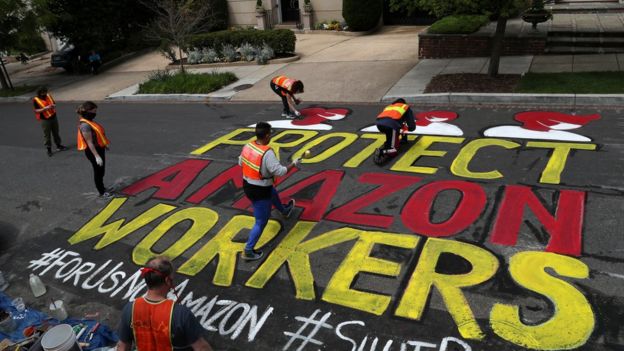 Several people contribute to paining a very large "protect Amazon workers" sign on the road outside a very affluent home