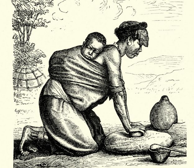 Mujer moliendo granos de rodillas, con un bebé en su espalda