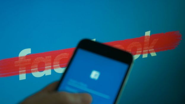 Celular con la aplicación de Facebook, delante de un logo de Facebook tachado.