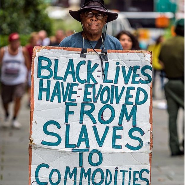 Activist holding sign reading BLACK LIVES HAVE EVOLVED FROM SLAVES TO COMMODITIES