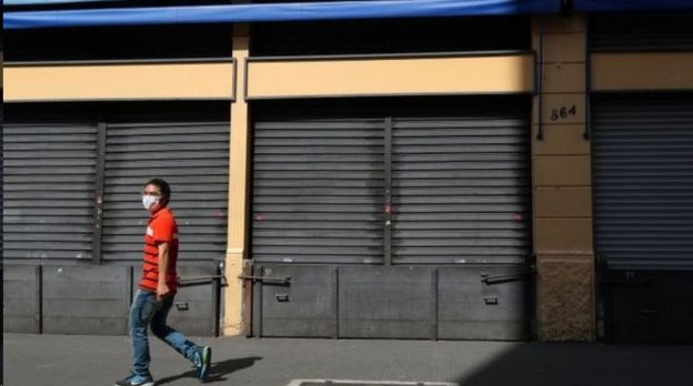 Homem caminha de máscara diante de lojas fechadas