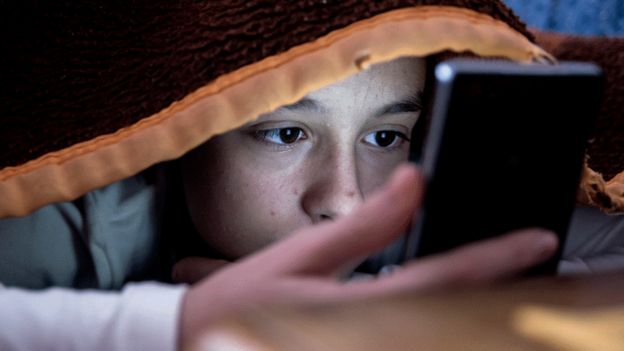 Foto mostra um menino debaixo de um coberto olhando para um celular; seu rosto está iluminado pela luz do celular