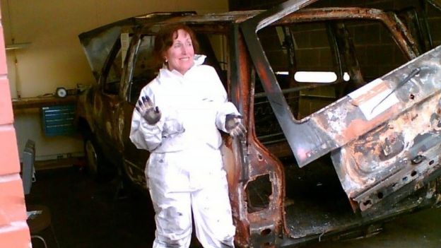 Wiltshire com jaleco, sorrindo e mostrando a mão suja de carvão ao lado de um carro queimado