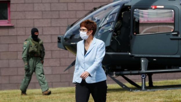De máscara, a ministra da defesa alemã, Annegret Kramp-Karrenbauer, caminha em pátio, com homem fardado e helicóptero atrás
