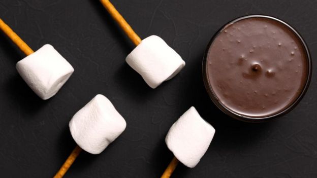 Palitos com marshmallow ao lado de pote de chocolate