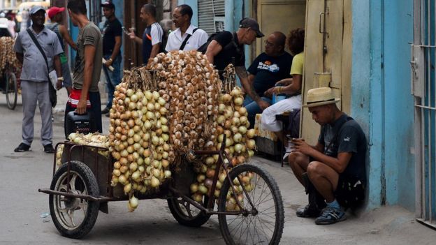 Imagem mostra pessoas em rua de Cuba onde também há um carrinho cheio de cebolas e alho, possivelmente à venda