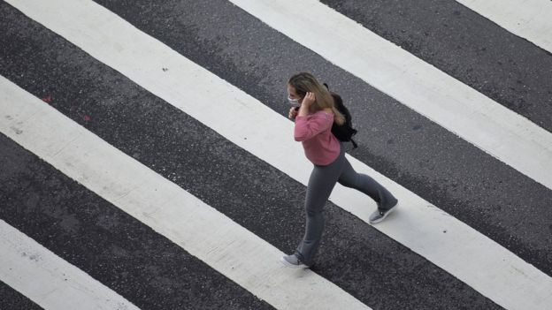 Em foto tirada do alto, mulher de máscara aparece atravessando faixa de pedestres