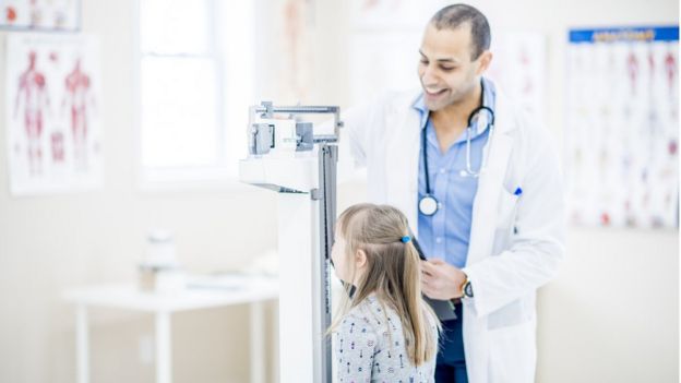 Menina com síndrome de Down em cima da balança em consultório, observada por um médico; ambos sorriem