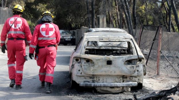Imagem mostra veículos incendiados e dois homens com uniformes da Cruz Vermelha caminhando na rua